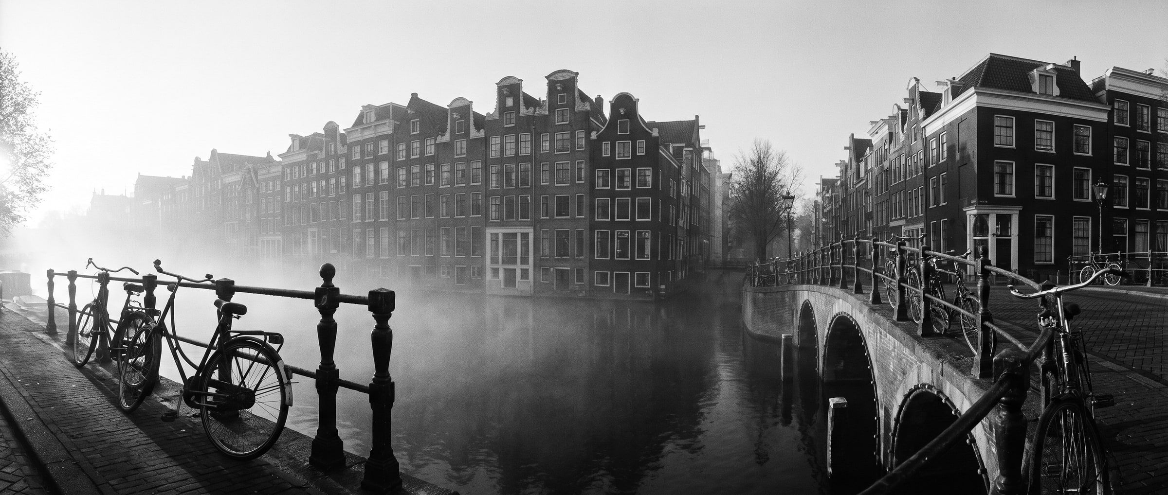 Amsterdamse grachtenpanden in de ochtendmist, met fietsen tegen een brugleuning