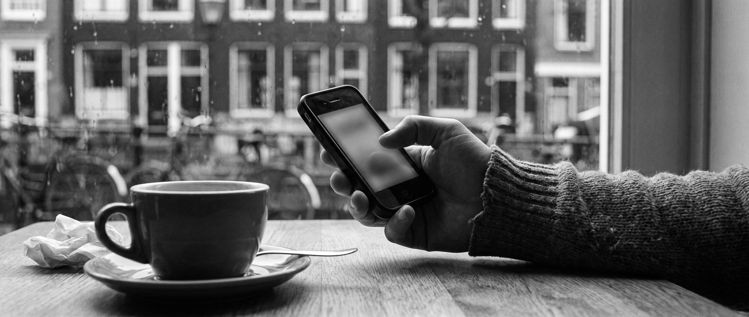Een hand met een telefoon aan een cafétafel, met Amsterdamse grachtenpanden op de achtergrond